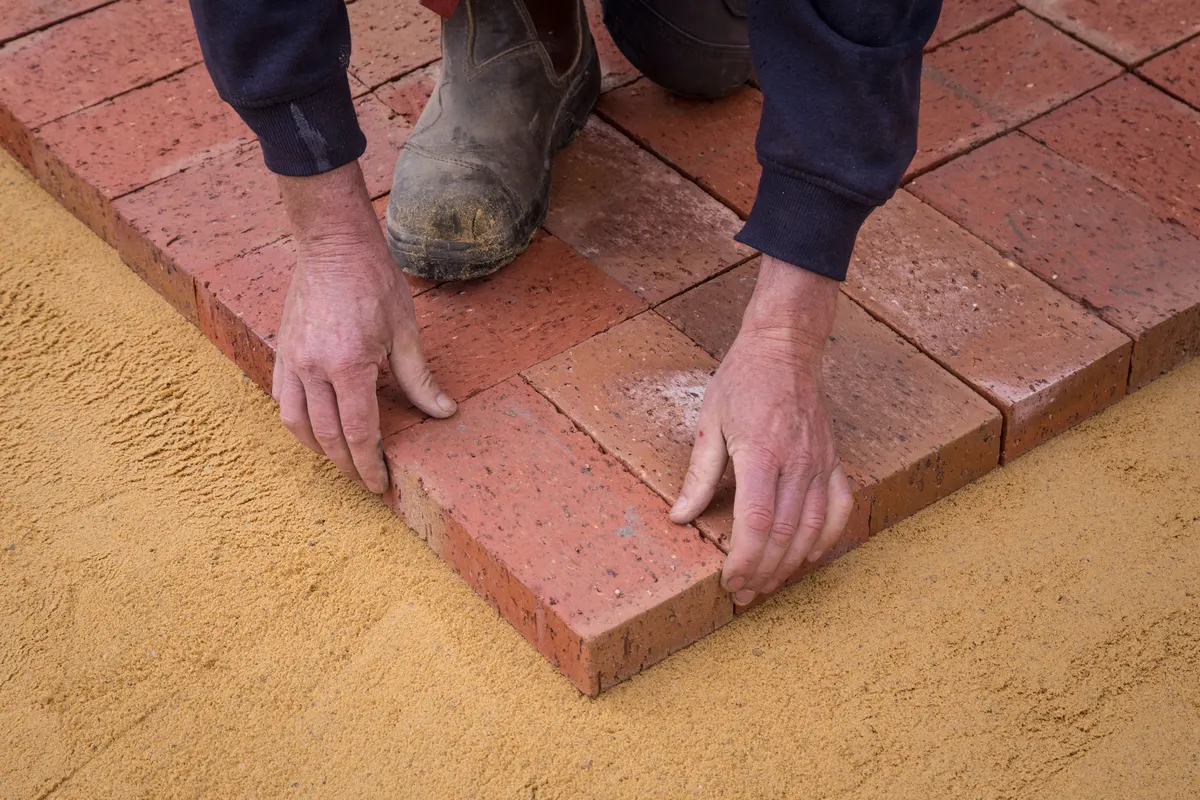 Hands setting a clay paver onto a screeded bedding sand layer